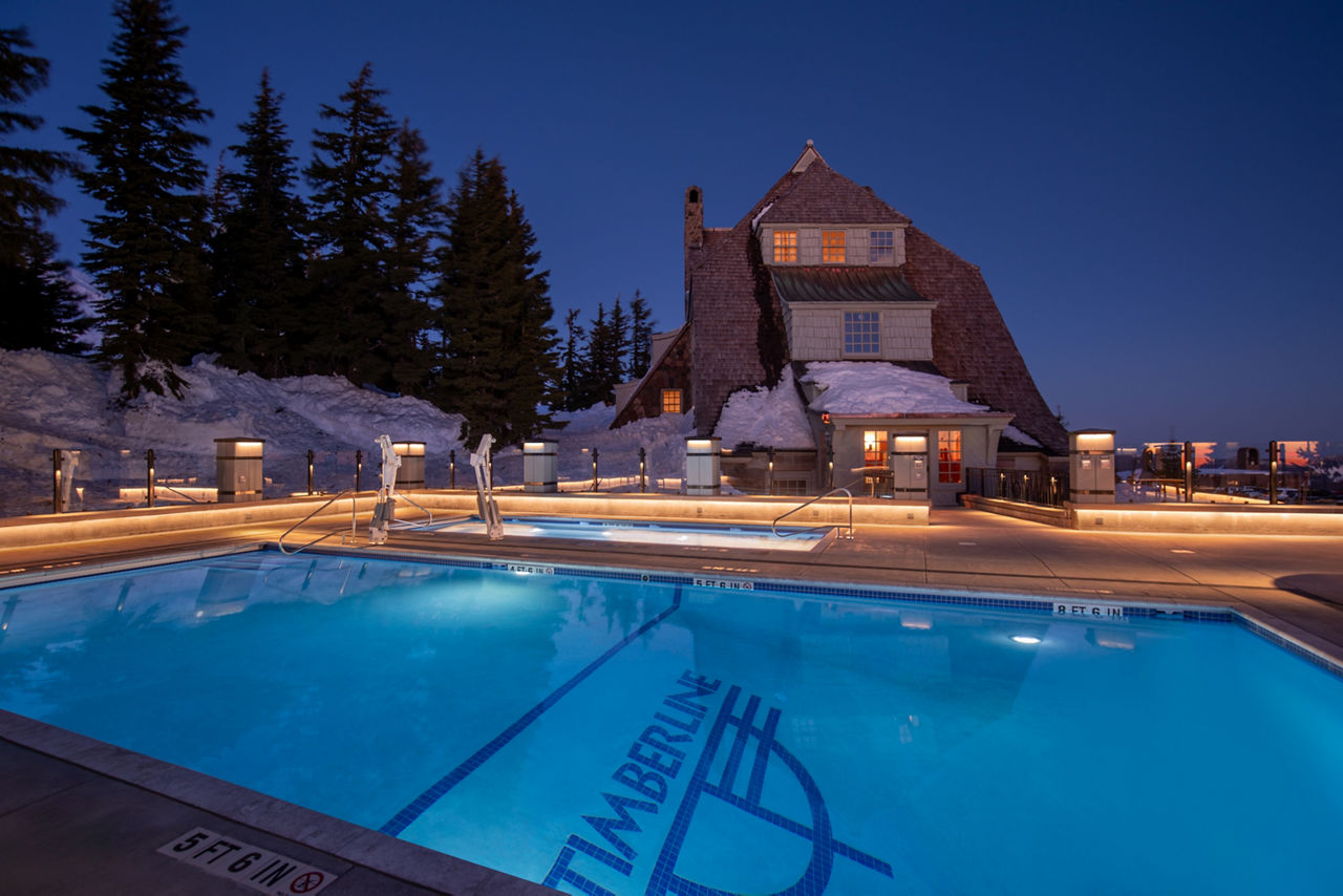 A glowing outdoor swimming pool is featured in front of a rustic mountain lodge at dusk
