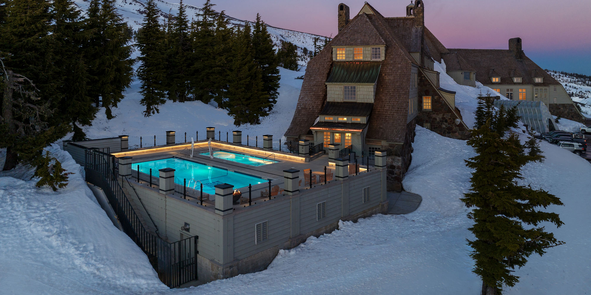 A large rustic lodge sits nestled in a snowy mountain landscape at dusk, with a glowing outdoor swimming pool surrounded by snow. 