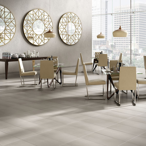Restaurant with gray concrete look rectangle floor tile, beige chairs, glass tables, gray walls, and floor to ceiling windows showing the city.