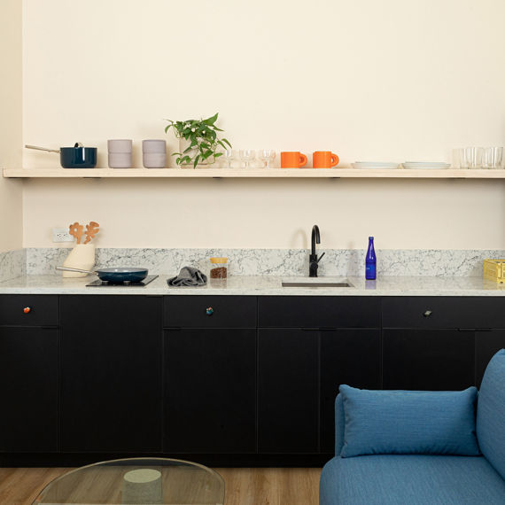 Kitchen with white quartz marble look countertop slab and backsplash, beige walls, shelf with plant and dishes, black cabinets, and white vase with wooden spoons.