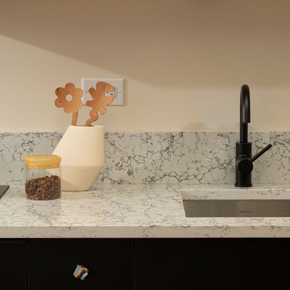 Kitchen with white quartz marble look countertop slab and backsplash, beige walls, shelf with plant, and vase with wooden spoons.