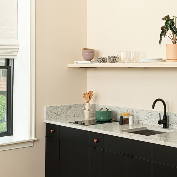 Kitchen with white quartz marble look countertop slab and backsplash, beige walls, shelf with plant and dishes, and white vase with wooden spoons.