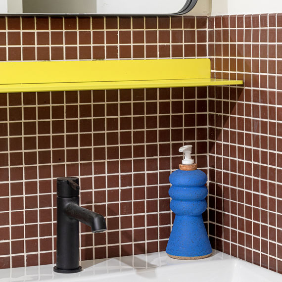 Close up of bathroom sink with brown square mosaic tile on backsplash, white sink, yellow shelf, and blue soap bottle.