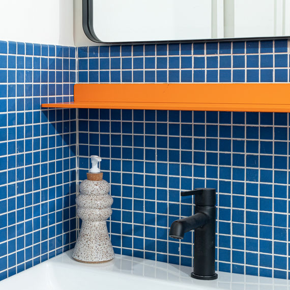 Close up of bathroom sink with deep blue square mosaic tile on backsplash, white sink, orange shelf, and gray soap bottle.
