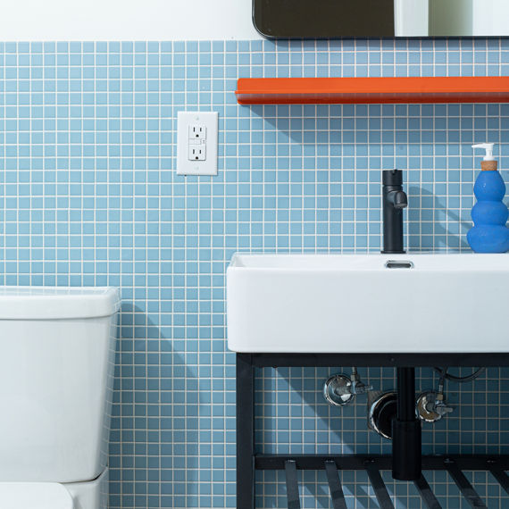 Close up of bathroom with light blue square mosaic tile on bottom half of wall, white sink, orange shelf, blue soap bottle, white top half of wall, and black accents.