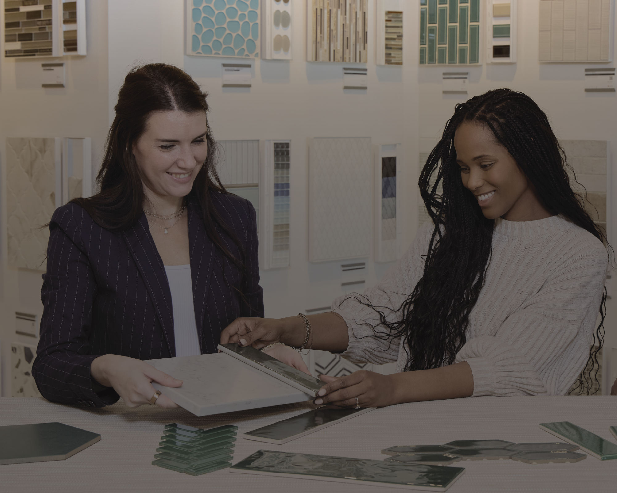 Two women sit at a table reviewing tile samples in a showroom. One person holds open a sample book while the other points to a specific tile. Various wall-mounted tile boards with different colors and patterns are displayed in the background. The sce