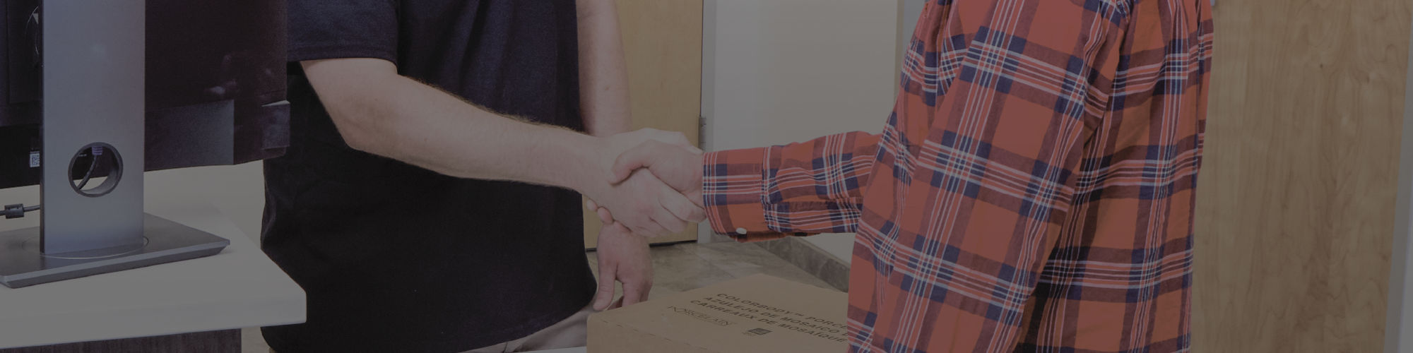 A customer in a plaid shirt shakes hands with an employee across a modern service counter. The employee stands behind a computer monitor and holds a flat cardboard box on the counter, suggesting a pickup or delivery transaction. Office doors and info