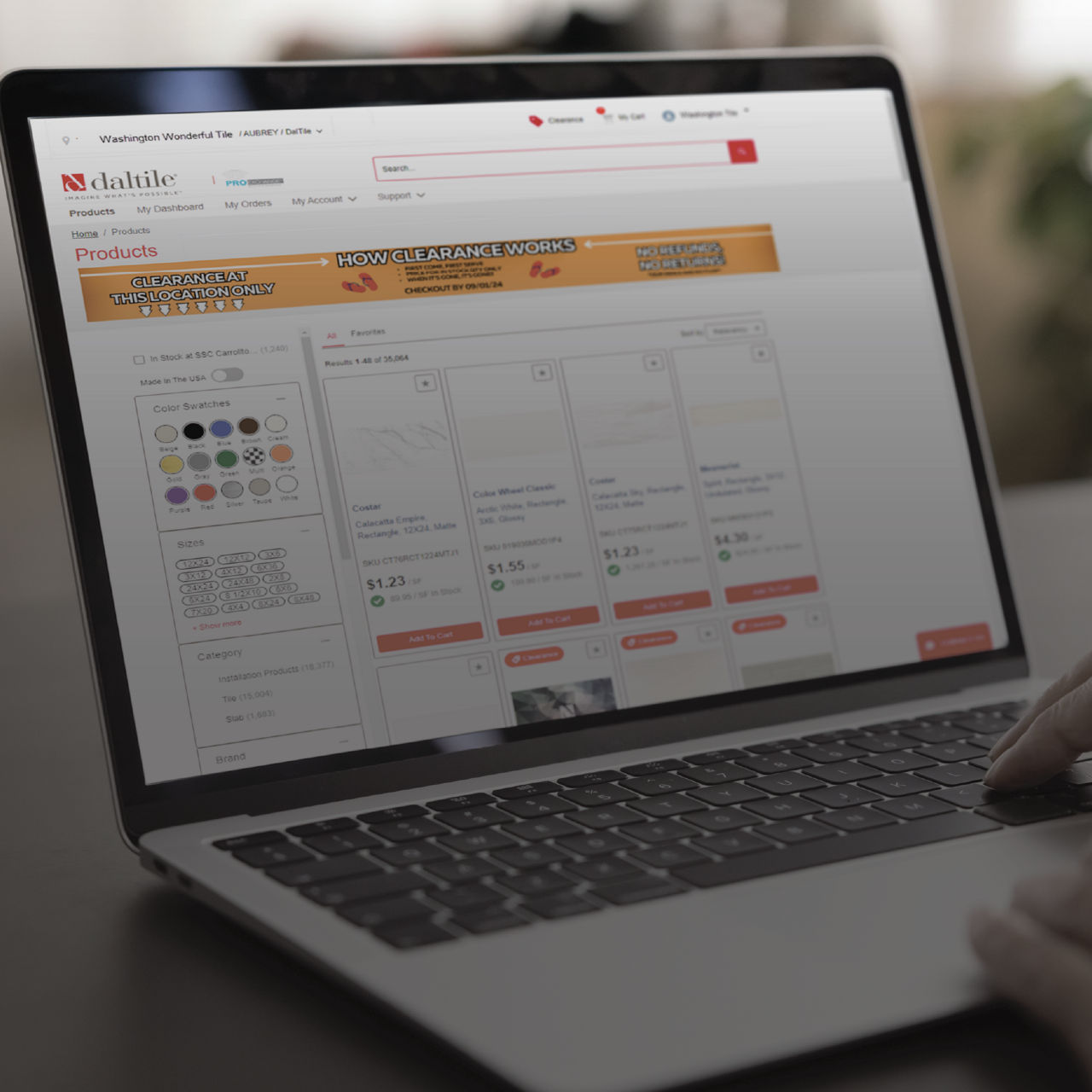 A person uses a laptop to browse an online catalog of tile products. The screen displays a website with the name "daltile" and several product cards showing prices and options. The scene is shot from an angle that focuses on the laptop screen and key