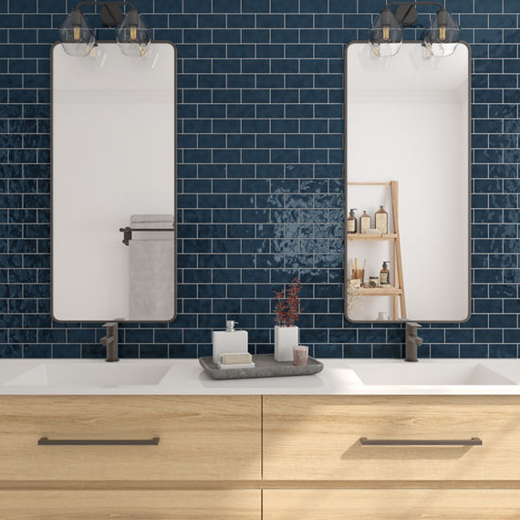 Bathroom with blue rectangle subway tile in brick joint pattern on back wall, light wood cabinets, white countertop, two large rectangle mirrors, and double sinks.