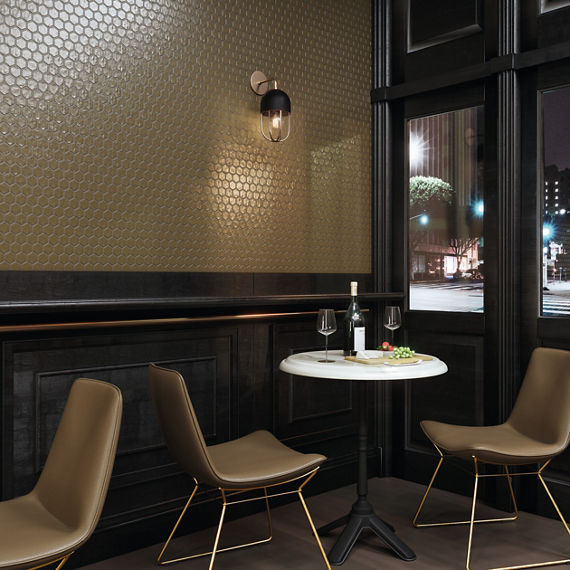 Restaurant with gold hexagon metal look mosaic tile on side wall, dark wood on back wall, small white table, and beige chairs.