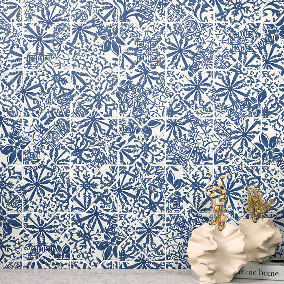 A decorative wall covered in blue and white floral patterned tiles forms the backdrop of this interior scene. In the foreground, two sculptural white ceramic vases sit on a smooth surface, holding dried grasses and stems. A book or box with the visib