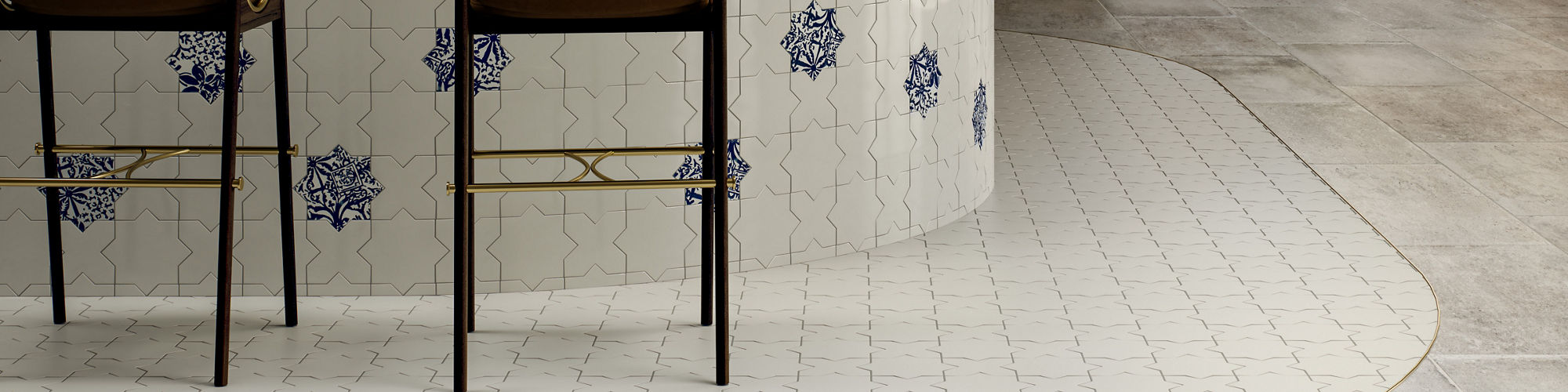 A stylish bar interior features a curved white tiled counter decorated with blue floral motifs. Two brown and gold bar stools sit at the counter beneath a suspended rack of hanging glassware. Backlit shelves filled with bottles line the wooden wall b