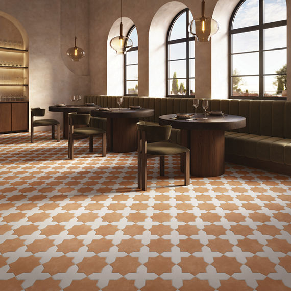 A contemporary restaurant interior features a striking geometric tile floor in warm terracotta and white tones. Round wooden tables with dark finishes are paired with cushioned chairs and a long green banquette along the wall. Large arched windows al