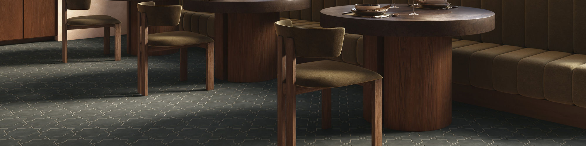 A stylish restaurant interior features round wooden tables and upholstered banquette seating along the wall. Large arched windows allow natural light to fill the space and reveal a view of trees outside. Pendant lights hang from the high ceiling, add