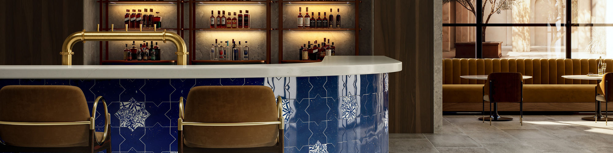 A contemporary bar interior features a curved counter clad in glossy blue tiles with white geometric patterns. Two upholstered bar stools with metal frames sit at the front of the counter, facing a back wall lined with illuminated shelves of bottles.