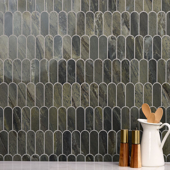 A modern tiled backsplash with elongated arch-shaped tiles forms the backdrop of this kitchen scene. The tiles feature a mix of dark green and gray tones with subtle natural stone veining. In the foreground, a white ceramic pitcher holds wooden cooki