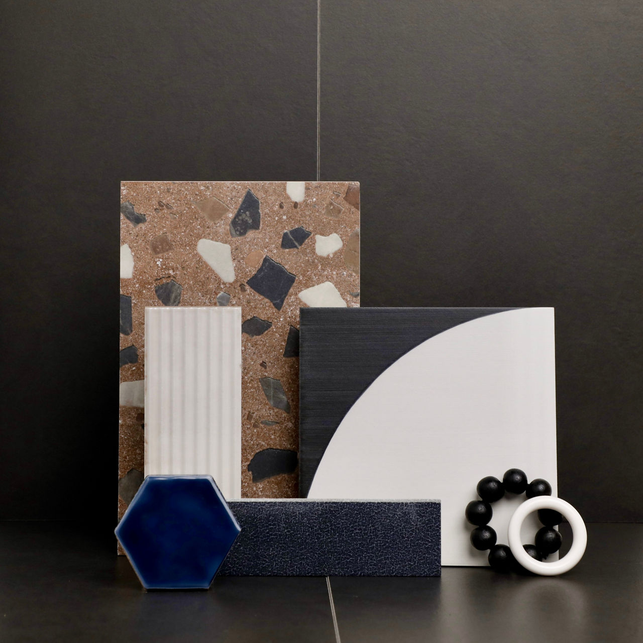 This flatlay contains a beige terrazzo look tile, black and white encaustic tile, black stone look rectangle tile, blue hexagon mosaic, white rectangle wall tile with linear design, and a black rectangle wall tile.