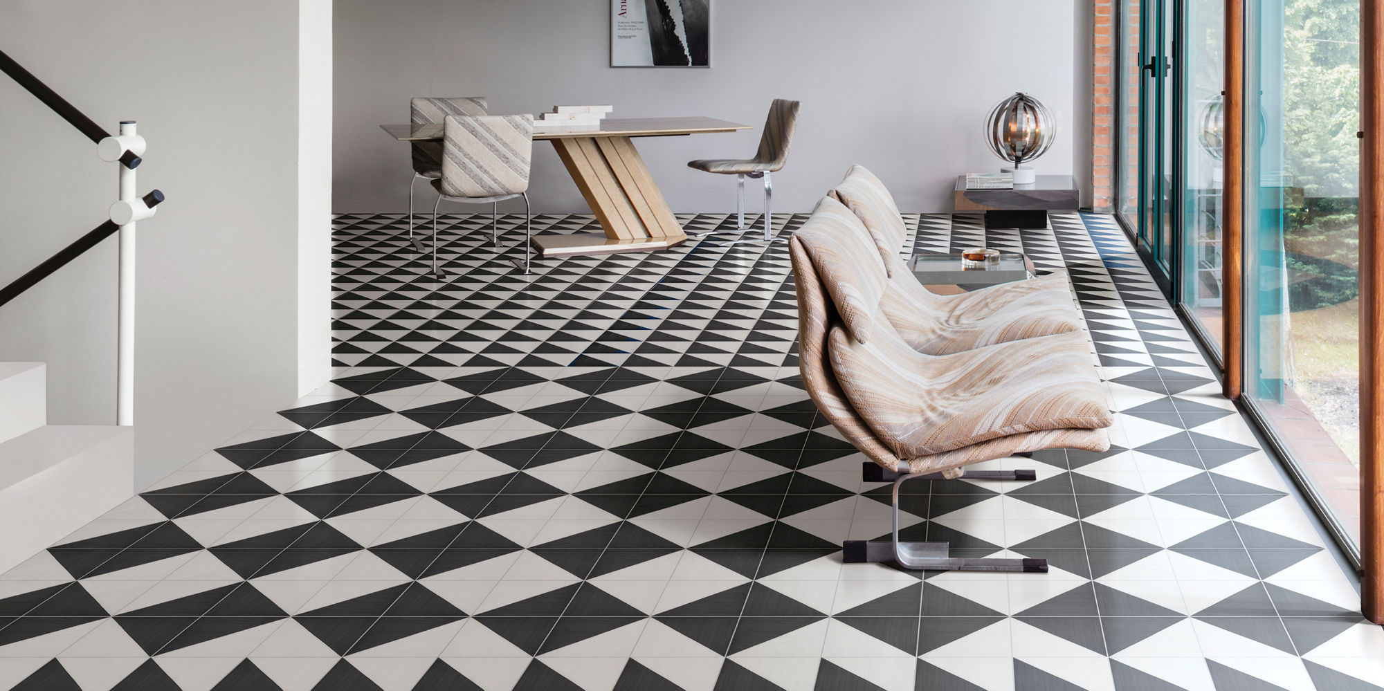 Contemporary open-plan interior featuring bold black and white geometric floor tiles. The space includes a minimalist staircase, a sleek wooden desk, and sculptural lounge chairs near tall glass windows. Natural light floods the room, highlighting th