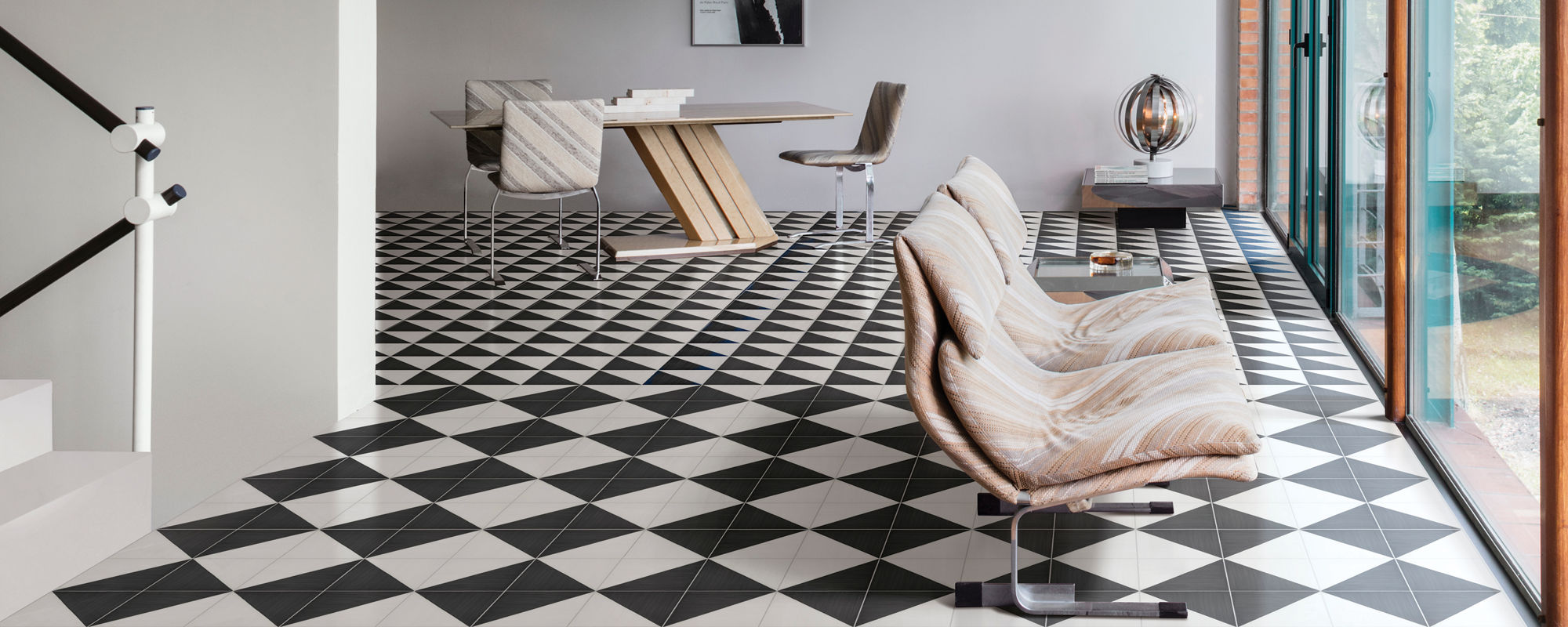 Contemporary open-plan interior featuring bold black and white geometric floor tiles. The space includes a minimalist staircase, a sleek wooden desk, and sculptural lounge chairs near tall glass windows. Natural light floods the room, highlighting th