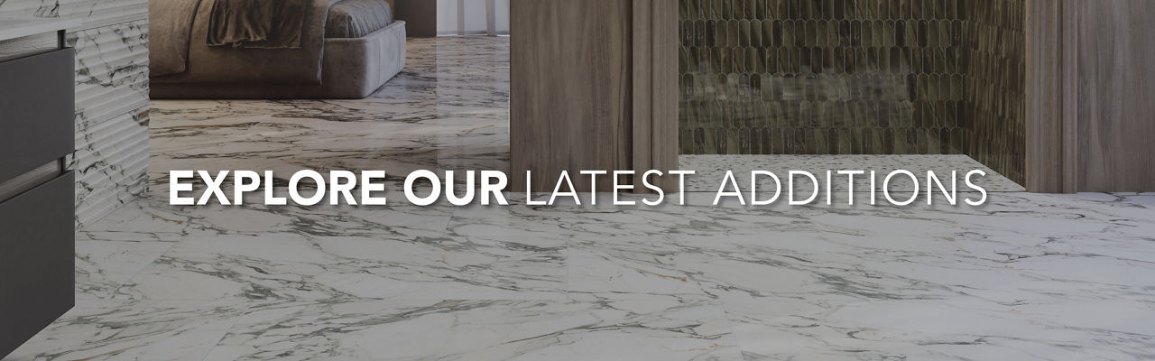 A contemporary interior space showcases sleek marble flooring in a neutral-toned room. Wooden cabinetry and a tiled wall add warmth and texture to the modern design. The text overlay reads "EXPLORE OUR LATEST ADDITIONS" in bold white letters across t