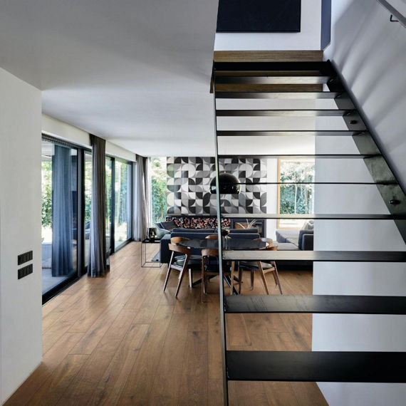 Open concept living room with stairs on side, wood floor planks, and floor to ceiling windows.