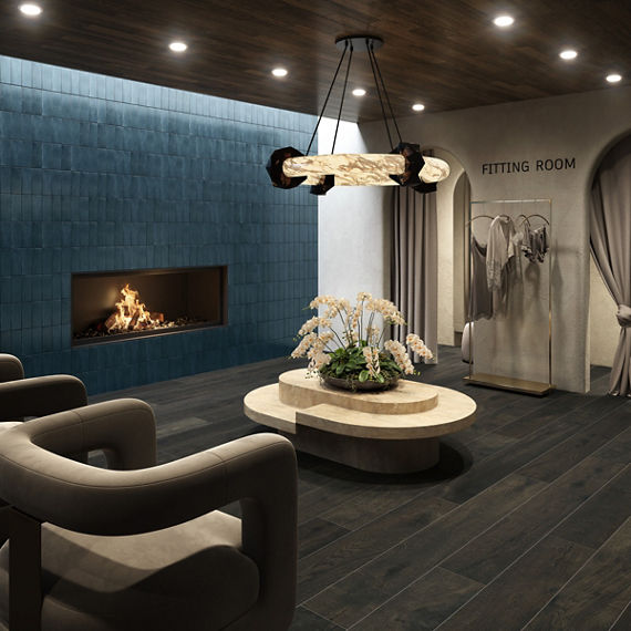 Fitting room waiting area with dark wood look floor tile, blue rectangle tile on side wall, fireplace, two gray chairs, and beige coffee table in center.