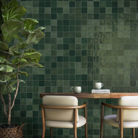 Dining area with dark green square wall tile, wood table, two beige chairs, two mugs and a stack of books on table, and tree on the side of the room.