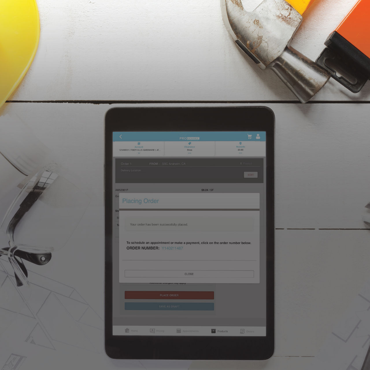 A digital tablet lies on a desk displaying an online order confirmation page. The interface shows an order number and options to place a check or print a receipt. Around the tablet are construction-related tools, including a hard hat and a wrench, su