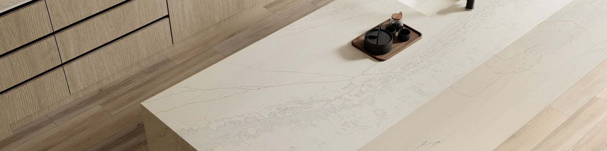 A spacious, contemporary kitchen features a large marble island with a sleek integrated sink and minimalist faucet. Light wood cabinetry and flooring create a warm, inviting atmosphere, complemented by neutral tones and subtle decor. The countertop i