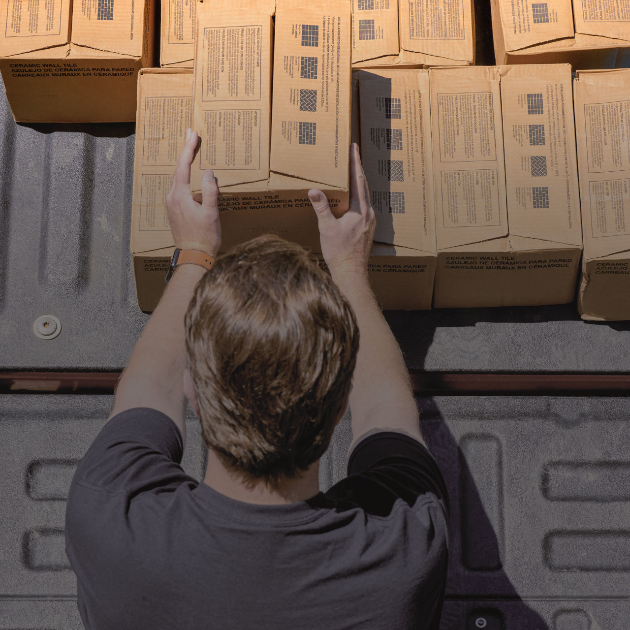 A person is seen from above arranging cardboard boxes in the back of a vehicle. The boxes are lined up in rows on a textured truck bed surface. Printed labels with text blocks and barcodes are visible on the top of each box, though the details are no