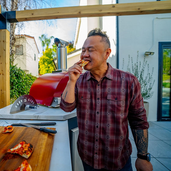 Jet Tila eating a bite of pizza in an outdoor kitchen with a beige quartzite countertop.