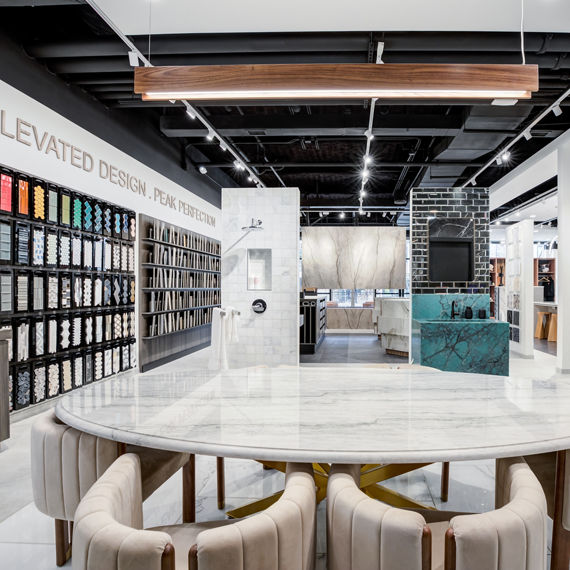 Modern tile showroom featuring a large marble table with upholstered chairs, wall displays of tile samples, and contemporary design elements in a bright, open space.