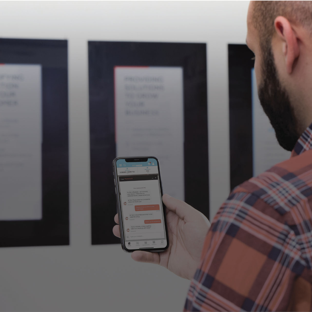 A person holds a smartphone while standing in front of wall-mounted displays. The phone screen shows a messaging or chat-style application interface with multiple text bubbles. The individual wears a plaid shirt and is viewed from behind in a modern 
