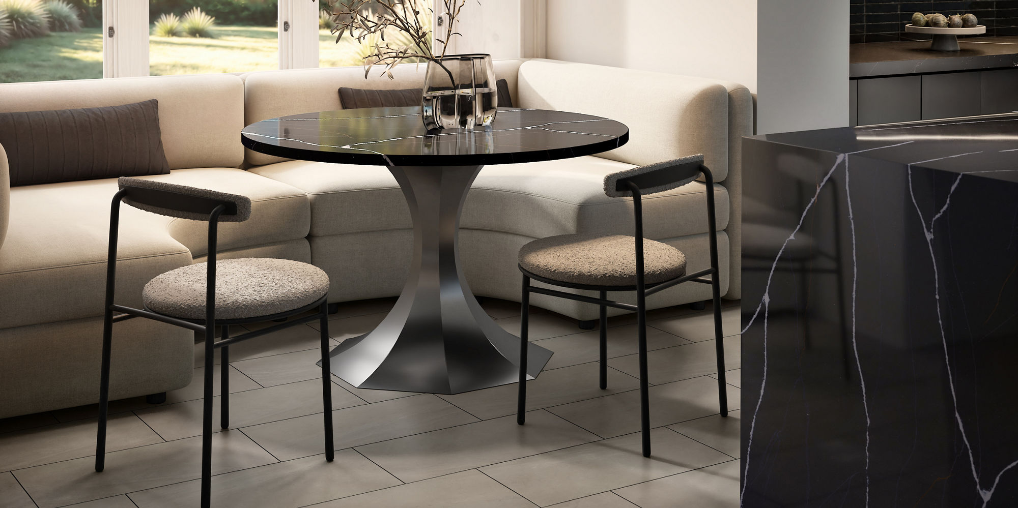 A contemporary dining nook features a sleek round table with a metallic pedestal base surrounded by minimalist chairs. The space is set against a large cream sectional sofa beneath tall windows that let in warm natural light and views of greenery out