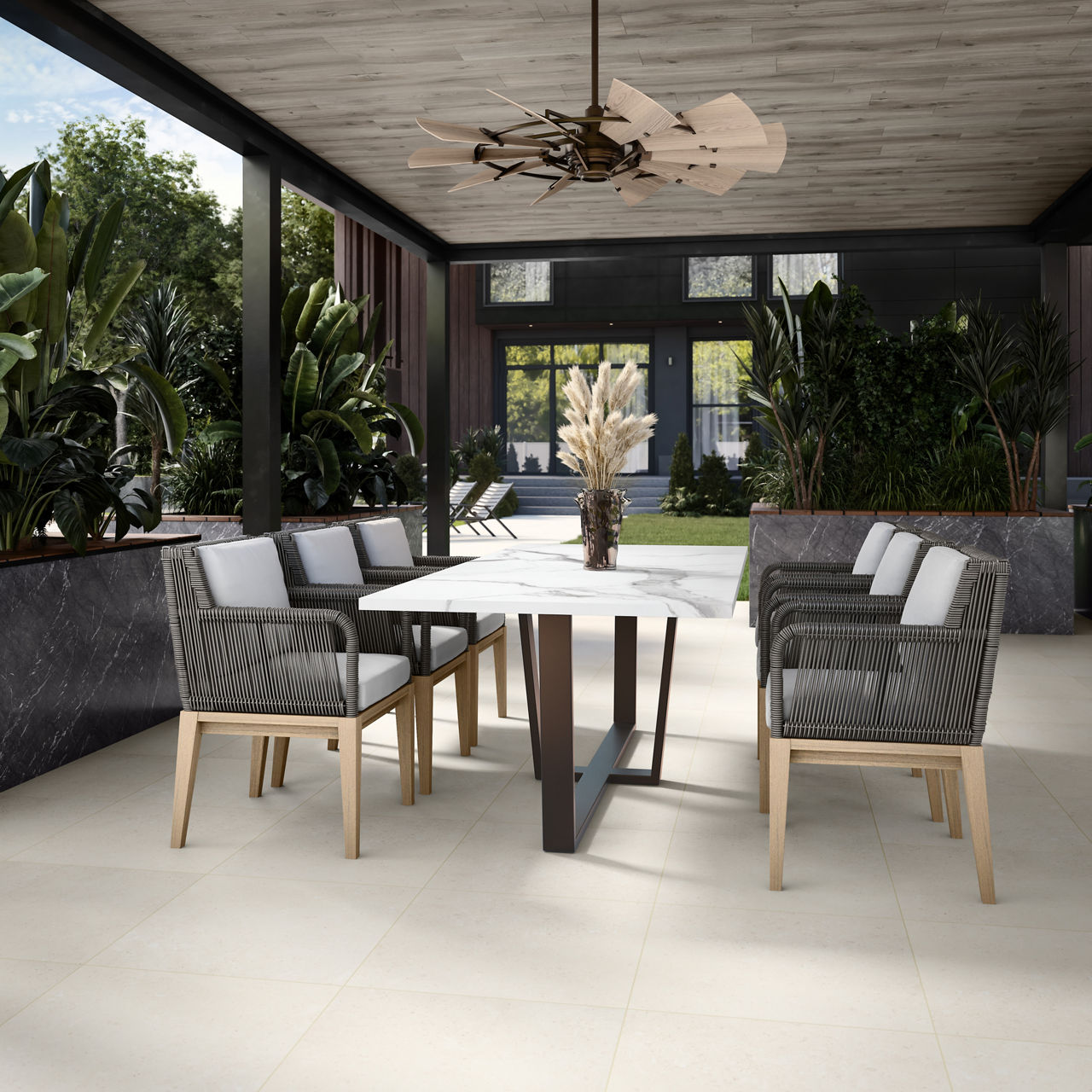 A contemporary outdoor dining space features a sleek rectangular table surrounded by cushioned armchairs. The area is set under a covered patio with a wood-paneled ceiling and a sculptural ceiling fan. Lush green plants and trees line the perimeter, 