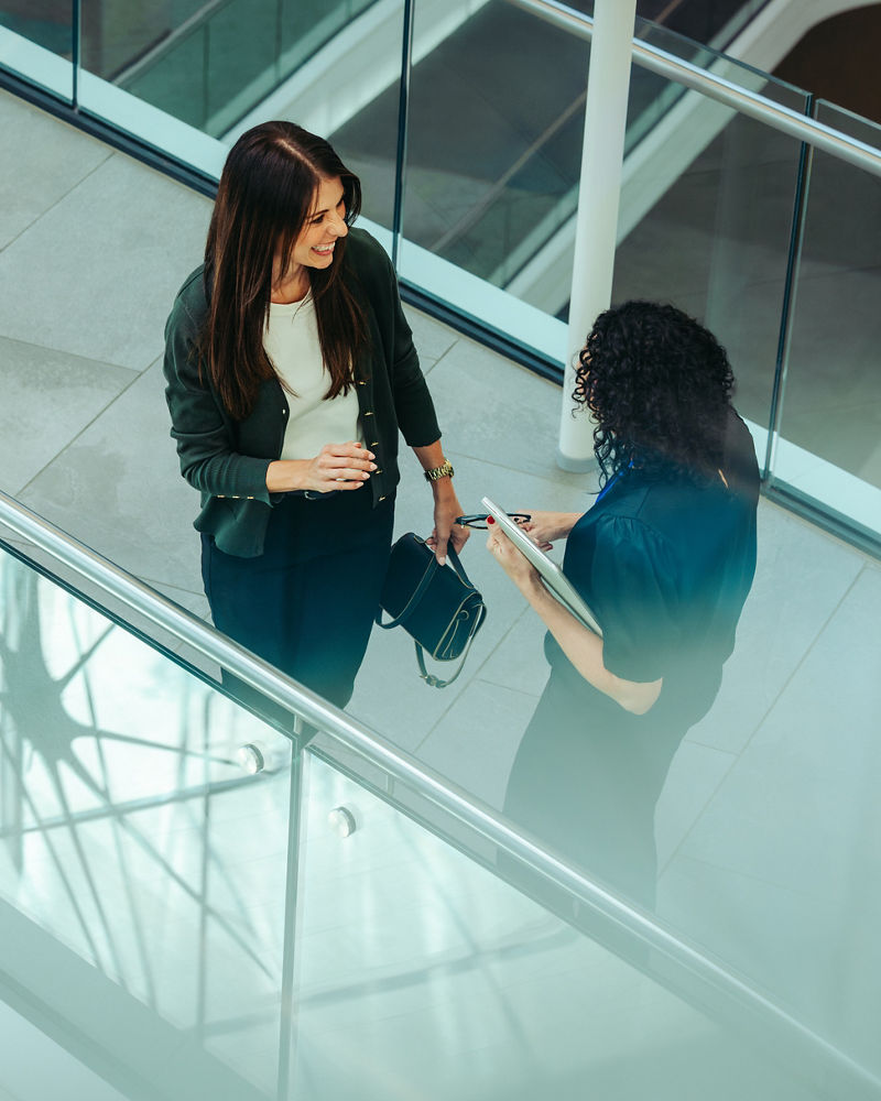 Two businesswomen engage in a friendly conversation while walking through a modern office building. They are discussing work-related topics, reflecting a productive and positive office culture.
