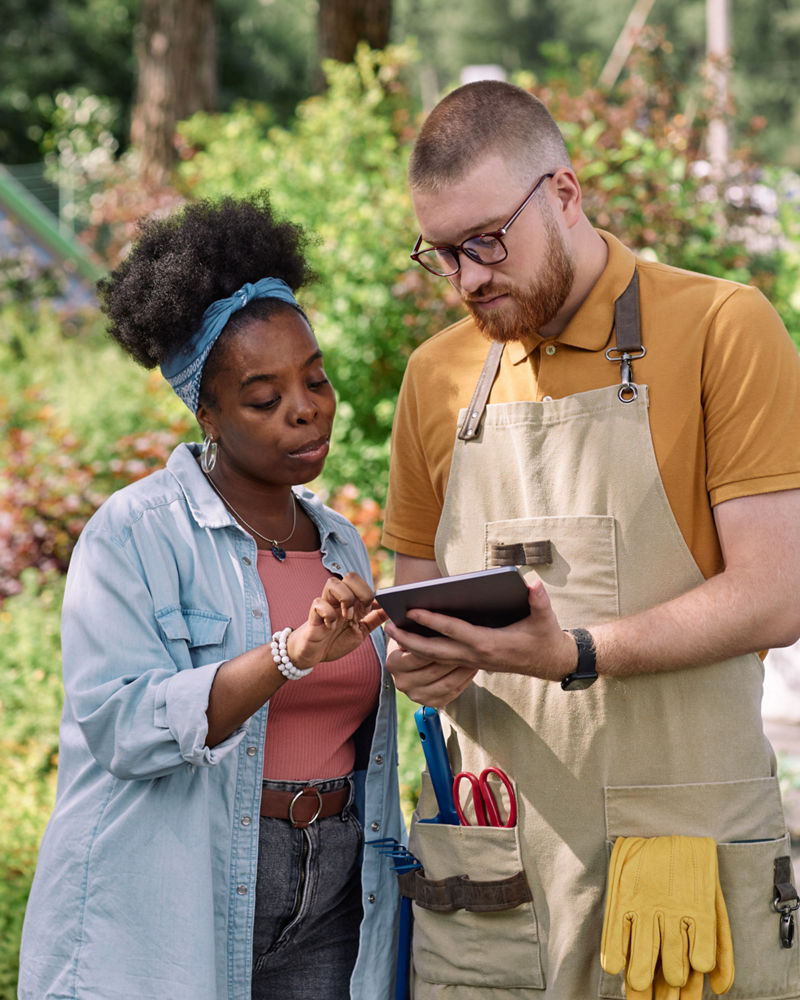 Woman pointing at tablet discussing project with plant nursery worker standing outside in sunlight