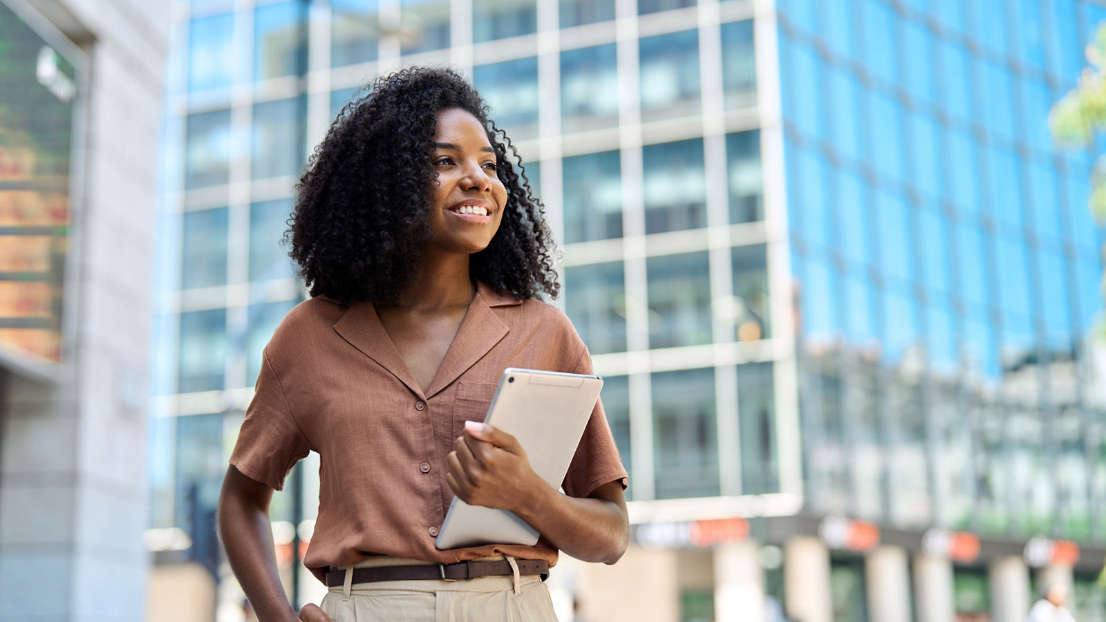 Happy confident young African American professional woman, student, female entrepreneur, businesswoman looking away holding tab standing on business city street dreaming of future career. Copy space.