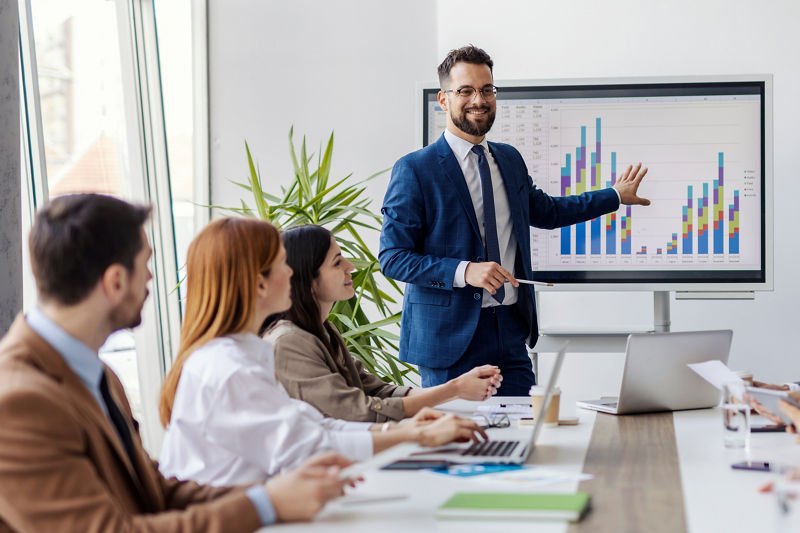 Portrait of smiling presenter standing near white board with statistics and explaining to his team financial report.