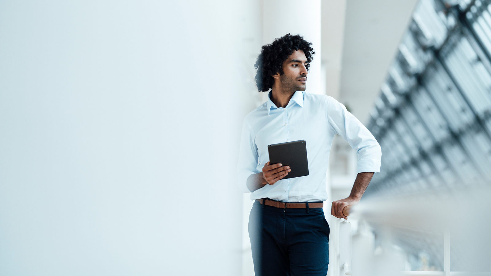 Thoughtful male professional holding digital tablet while standing by railing looking away in corridor at office