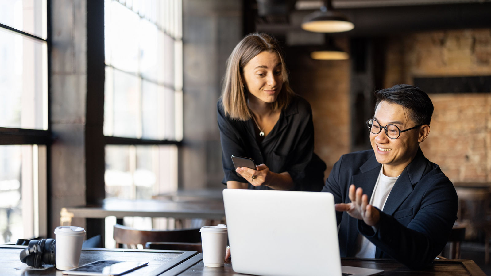 Asian businessman showing something on laptop computer to caucasian businesswoman in cafe. Concept of remote and freelance work. Idea of teamwork and business cooperation. Modern interior at sunny day