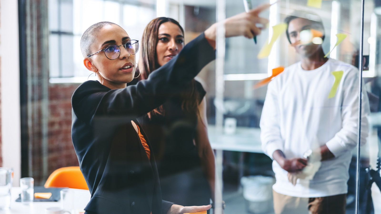 Smart businesswoman sharing her ideas with her team. Confident young businesswoman sharing her sticky notes with her colleagues while standing in front of a glass wall. Businesspeople working together.