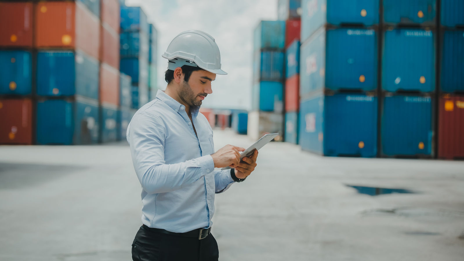 Engineer worker in causual suit standing in shipping container yard holding laptop with smile. Import and export product. Manufacturing transportation and global business concept