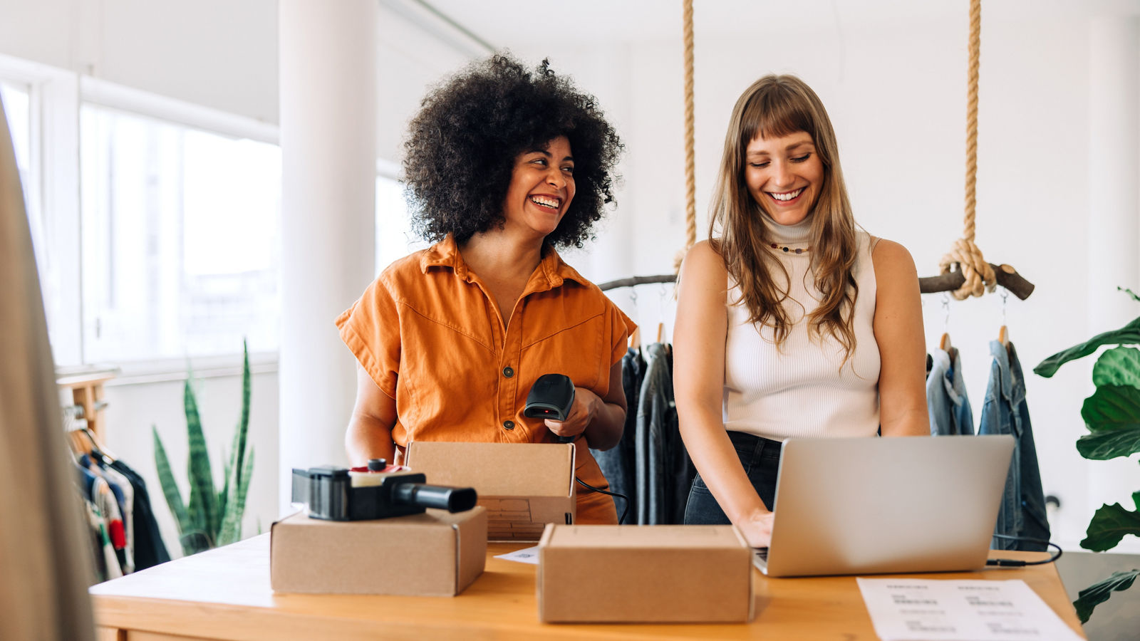 Cheerful clothing store owners preparing online orders for shipping. Two happy businesswomen smiling while working together in a thrift store. Female entrepreneurs running an e-commerce small business.