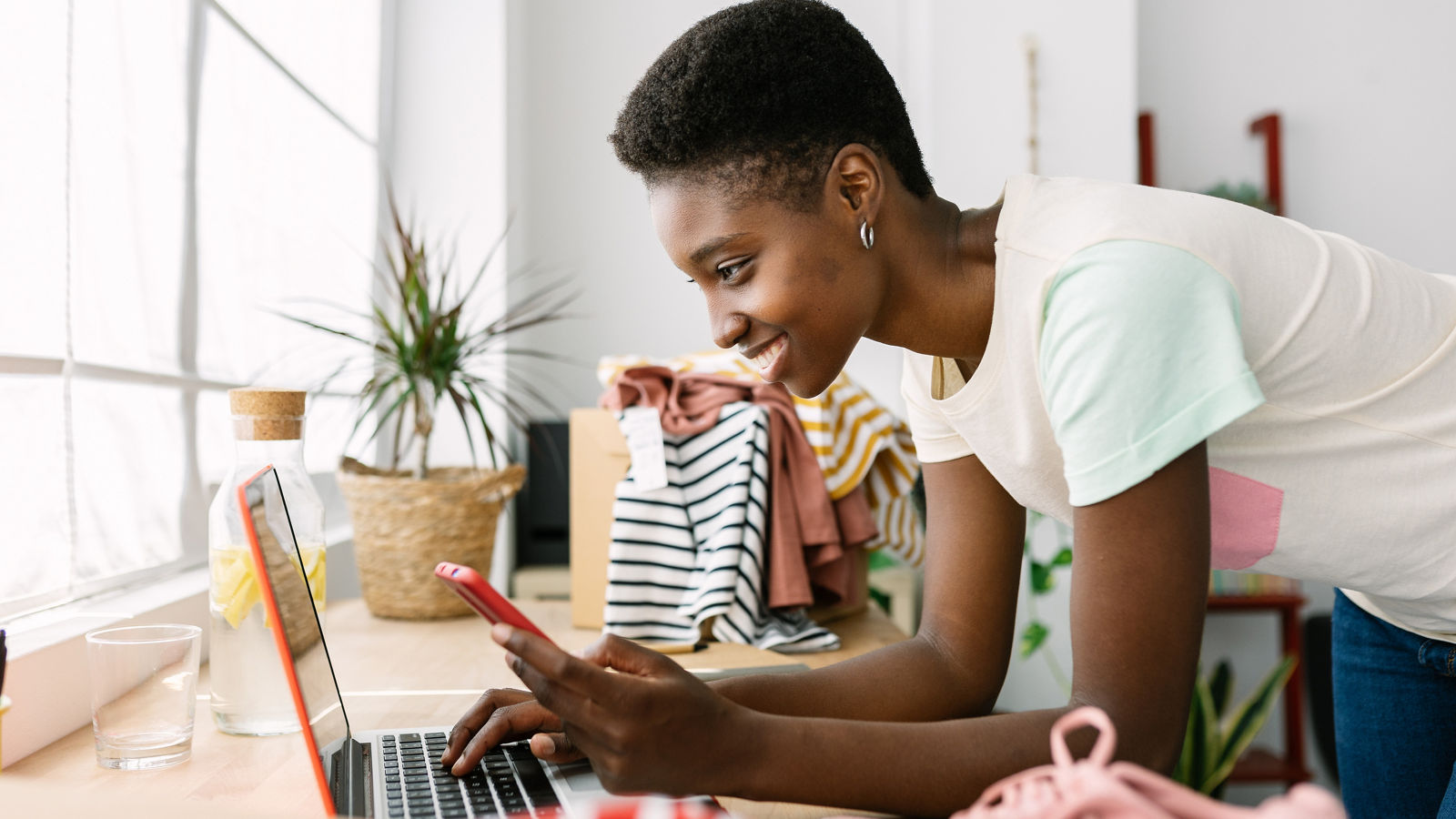 Small business owner woman looking at phone and laptop in the middle of her clothing inventory