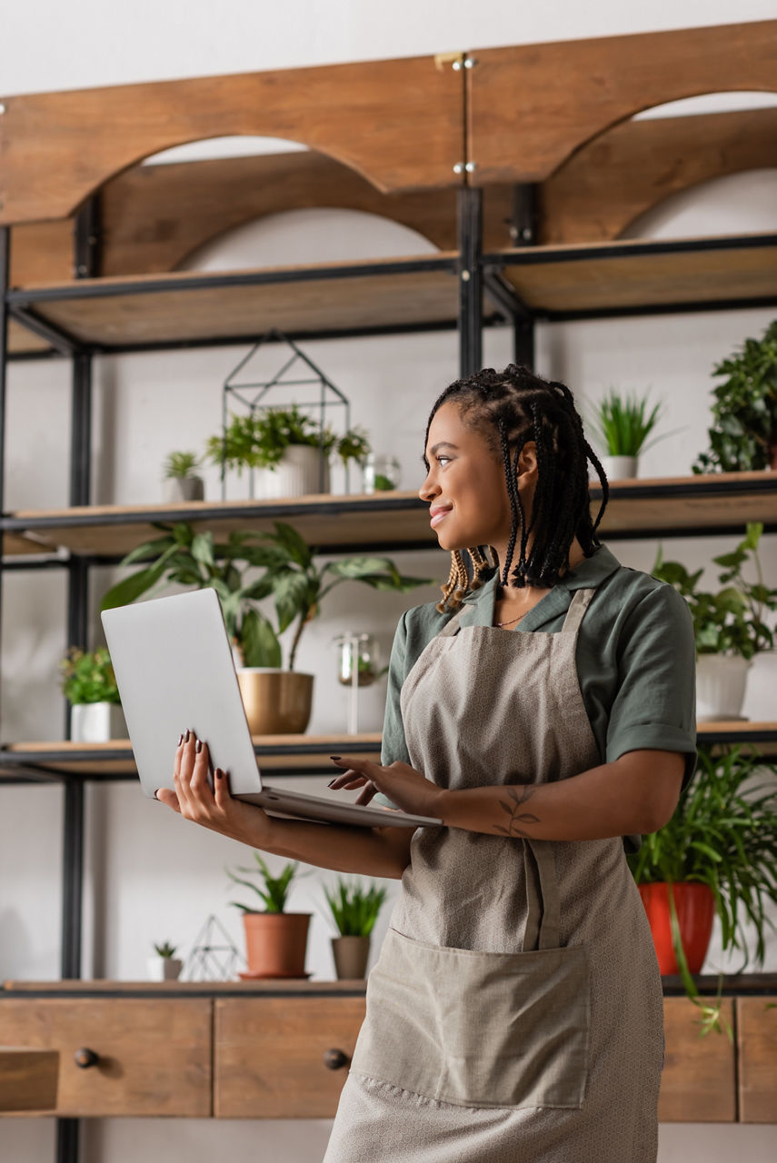 happy african american florist in apron holding laptop and looking away in flower shop