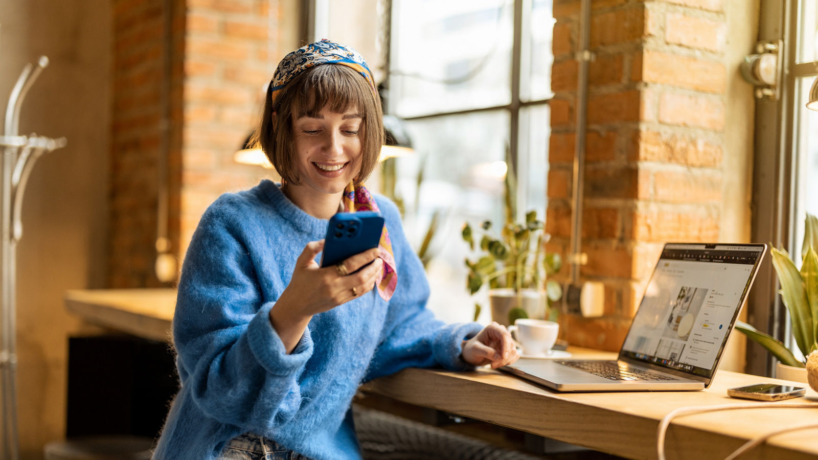 Young cheerful woman works on phone and laptop while sitting by the window with a coffee drink at modern cafe. Concept of remote work from public place, digital freelance and modern lifestyle