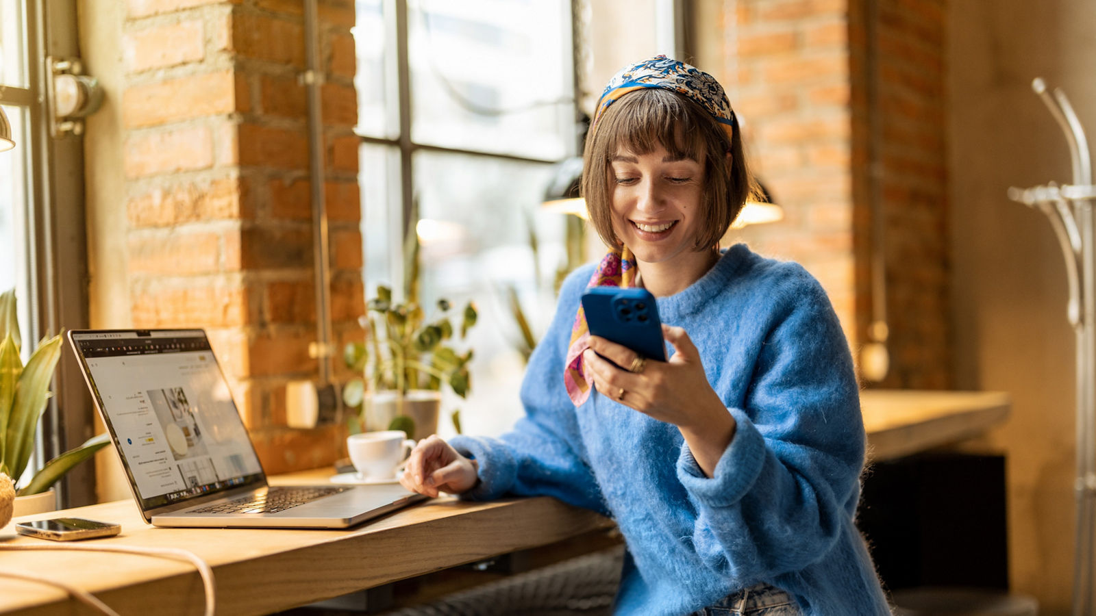 Young cheerful woman works on phone and laptop while sitting by the window with a coffee drink at modern cafe. Concept of remote work from public place, digital freelance and modern lifestyle