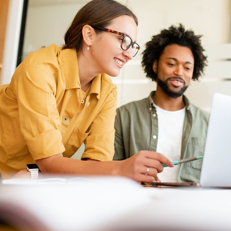 Two colleagues have pleasant conversation sitting side by side in modern open space office in front of laptop, smiling and enjoying friendly working atmosphere, brainstorming together, solving tasks