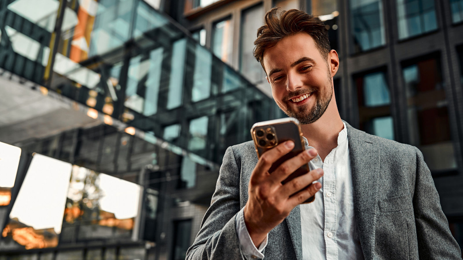 Portrait of attractive confident modern smiling happy adult dressed gray suit man holding a phone and standing outside near office building. Online work, communication. Copy space.                               